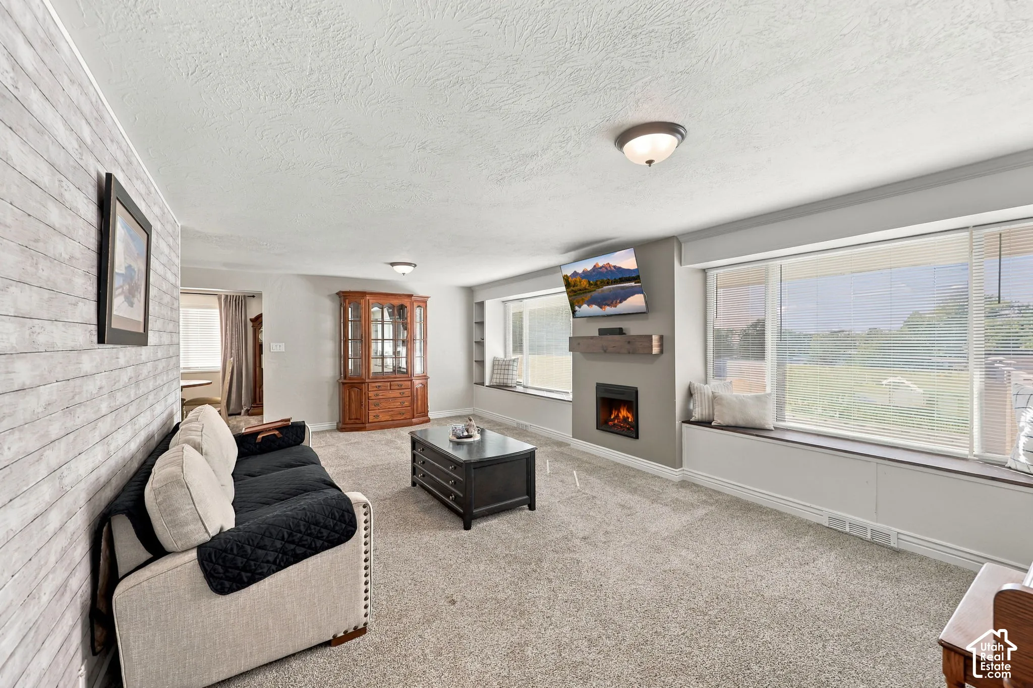 Electric fireplace with a wood-finish mantel, broad window sills, textured accent wall, and light-toned carpeting