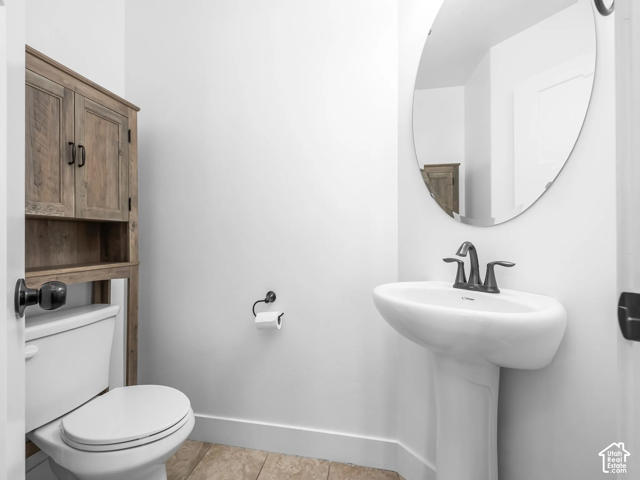 Pedestal sink with dark-finished faucet, oval wall-mounted mirror, and wood-finish storage cabinet