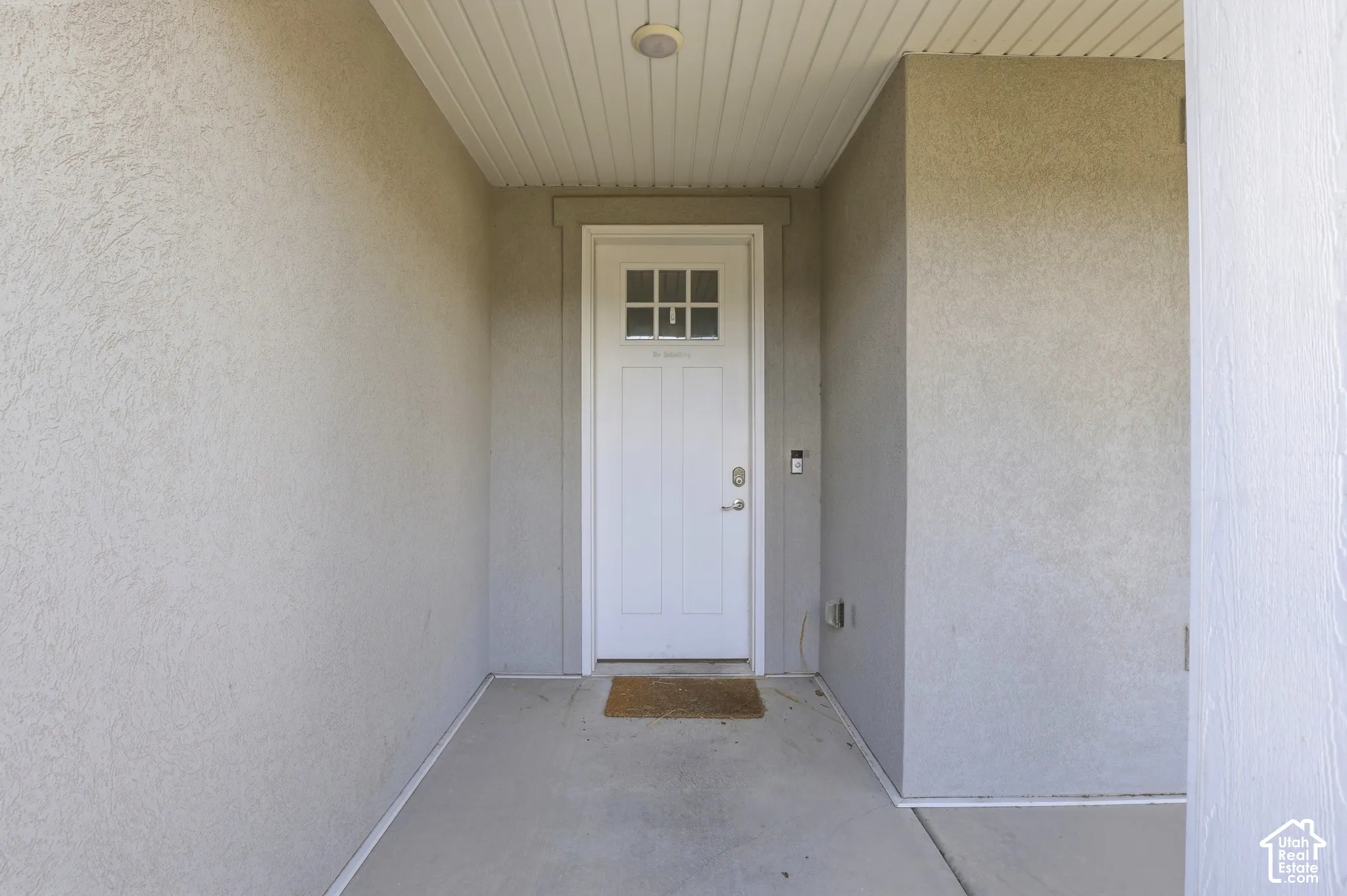 Recessed entry with stucco walls, a paneled white door with upper window panes, and a beadboard ceiling