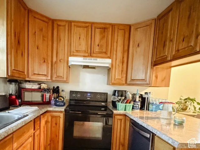 Wood-finish cabinetry, black range, integrated dishwasher, light-toned countertops, and rectangular tile flooring
