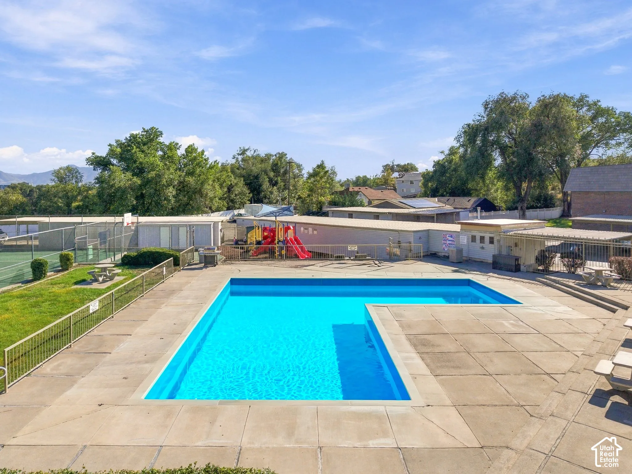 Community swimming pool with concrete decking, surrounding green space, and visible tennis court