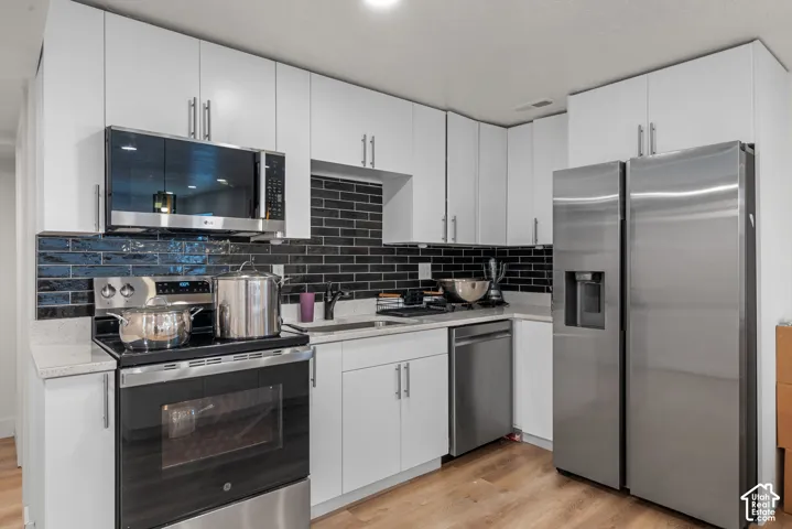 Kitchen featuring white upper and lower cabinetry, stainless steel appliances, black subway tile backsplash, light-toned countertops, and wood-finish flooring