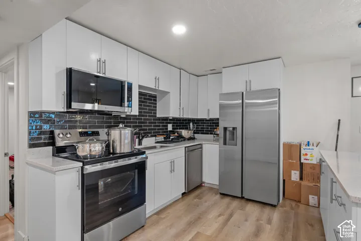 Modern kitchen featuring white cabinetry, stainless steel appliances, a black subway tile backsplash, and light wood-finish flooring