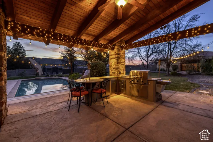 Covered patio with exposed wood beams, outdoor kitchen featuring stainless steel appliances, stamped concrete patio, stone-clad pillar, and an in-ground swimming pool
