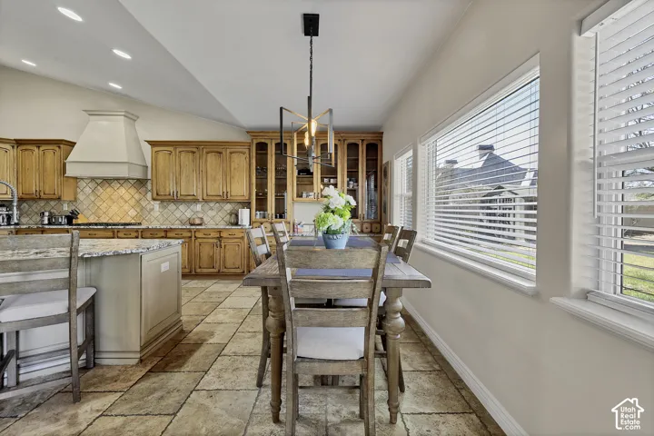 Sophisticated kitchen and dining area featuring custom wood cabinetry with decorative detailing, tile flooring throughout, stainless steel appliances, a pendant light fixture with brass accents, and expansive windows with plantation shutters providing nat