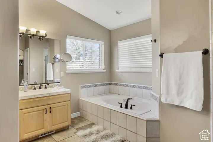 Built-in corner bathtub with tiled surround, wood-finish vanity with countertop, wall-mounted light fixture, and window with blinds