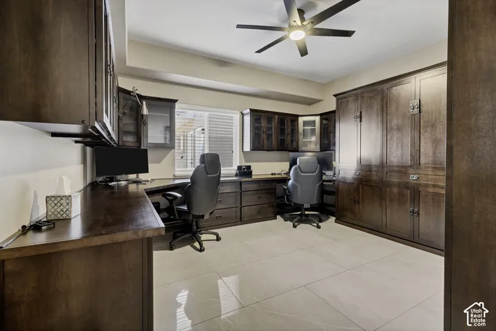 Spacious home office featuring dual workstations with dark wood cabinetry, polished tile flooring, recessed ceiling with ceiling fan, and abundant natural light from large windows