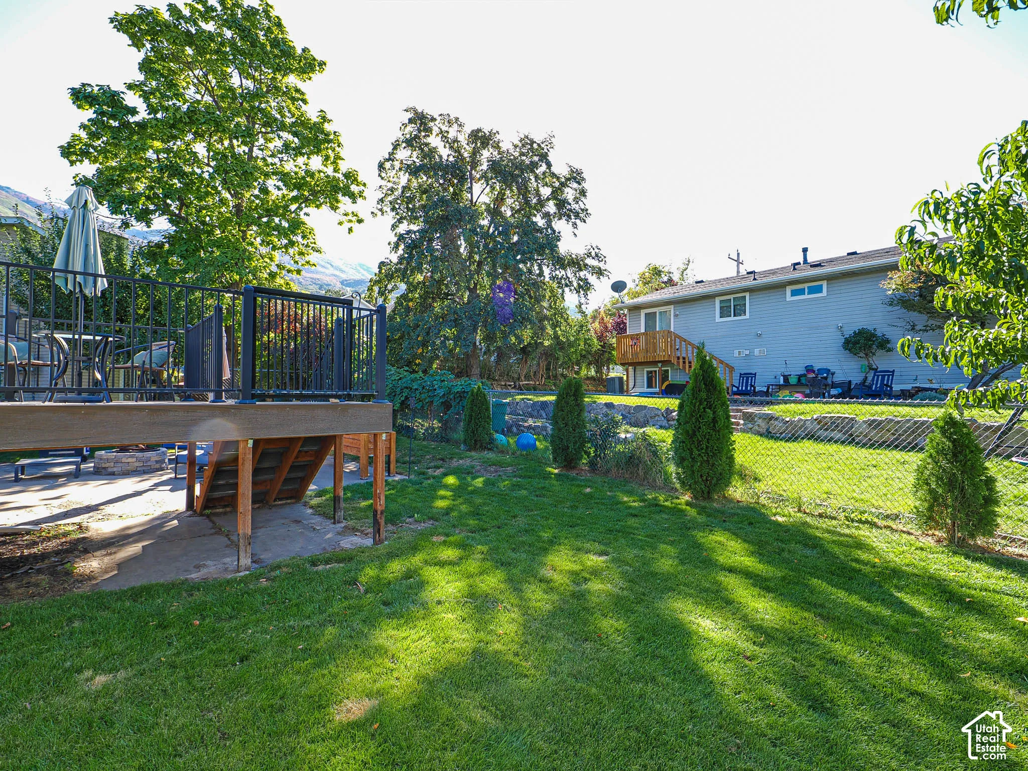 Expansive backyard featuring a green lawn, chain link fencing, a raised wood deck with black metal railing, and mature trees