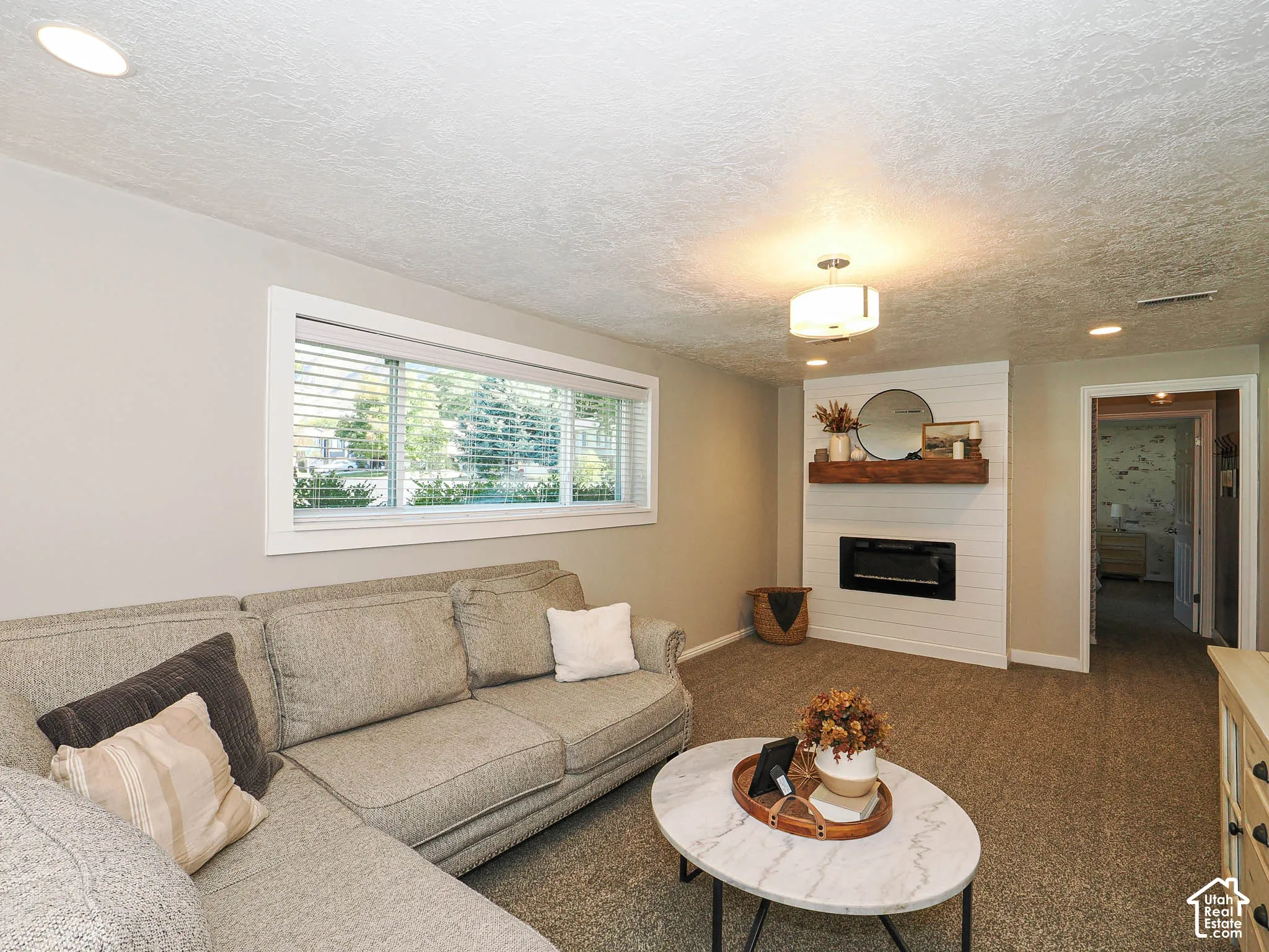 Recessed lighting, large horizontal window, shiplap-accented fireplace surround, wood-finish mantel, and carpet flooring