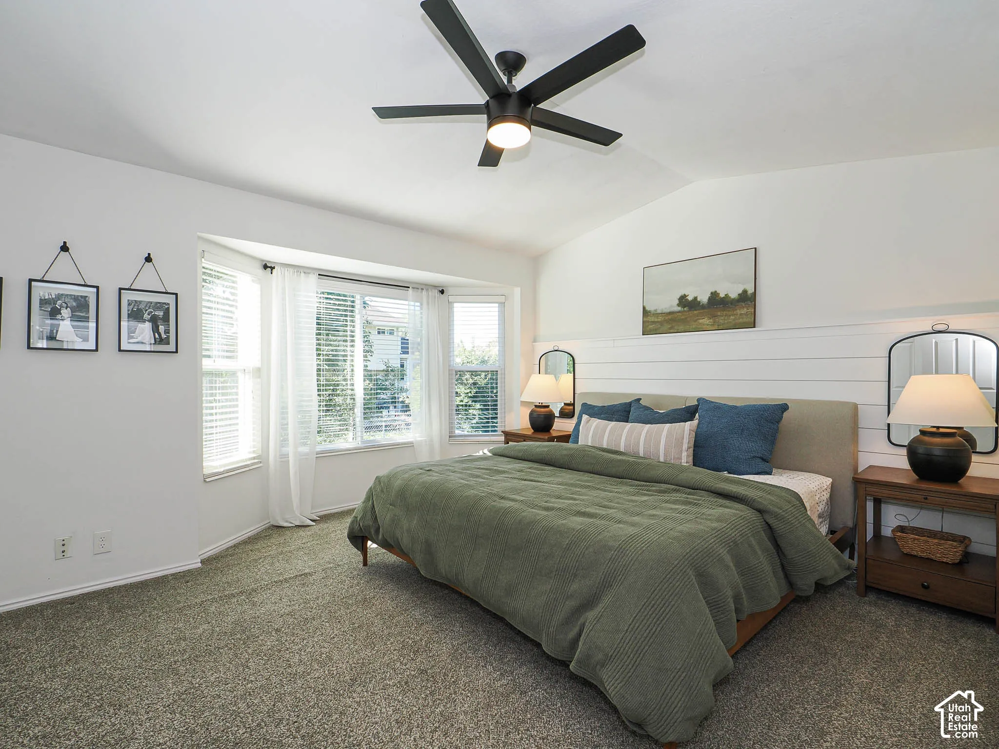 Vaulted ceiling, bay window, shiplap accent wall, dark-finish ceiling fan, and gray carpet