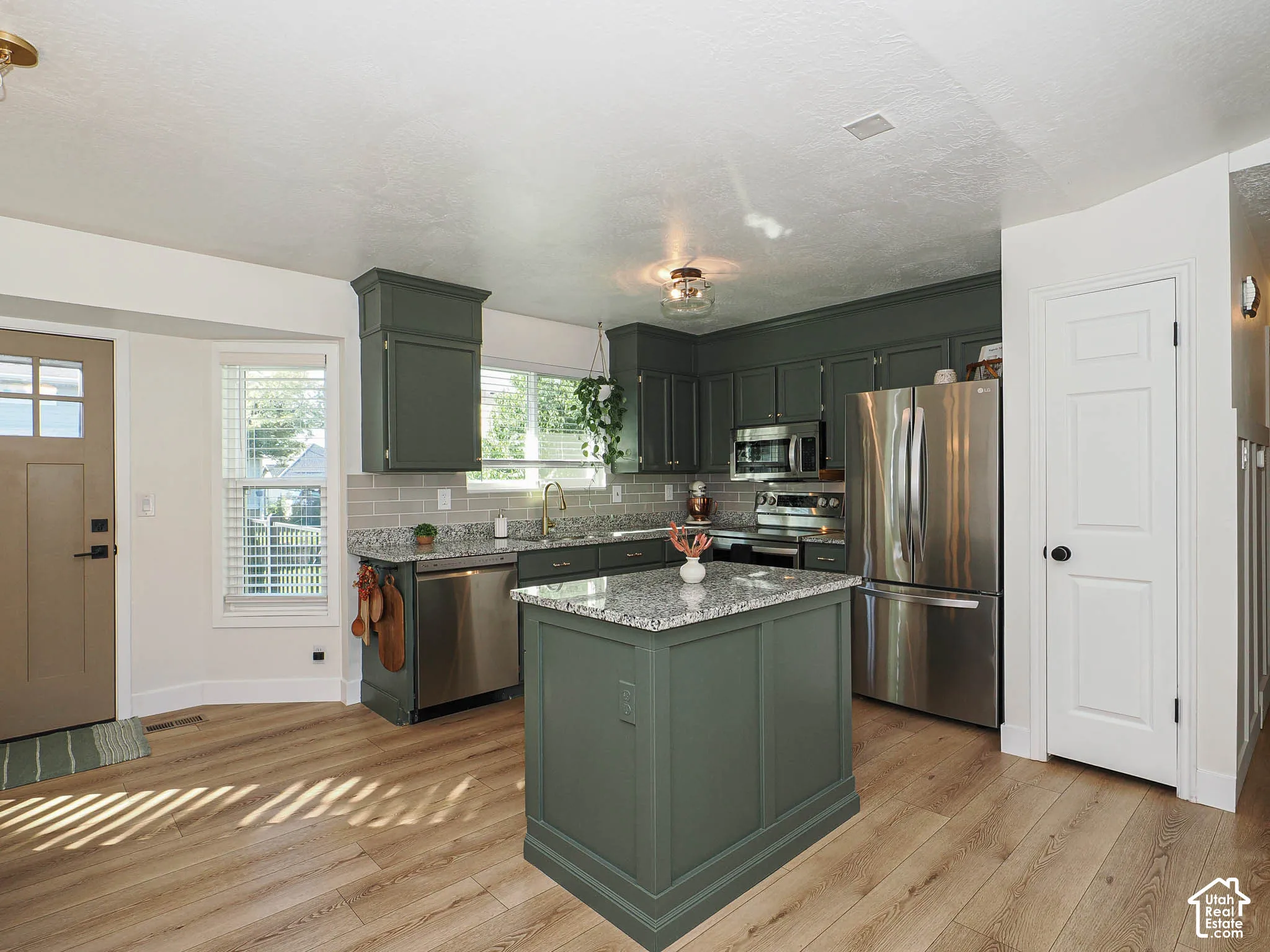 Kitchen featuring wood-finish flooring, dark green cabinetry, a central island with a light-toned countertop, stainless steel appliances, and a tiled backsplash