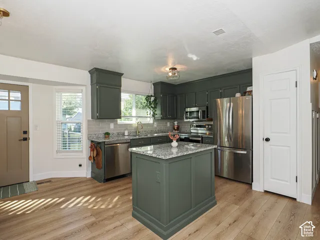 Kitchen featuring wood-finish flooring, dark green cabinetry, a central island with a light-toned countertop, stainless steel appliances, and a tiled backsplash
