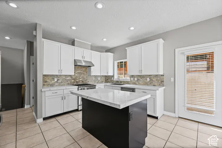 White shaker cabinetry, stone-look backsplash, stainless steel range with matching hood, central island with stone countertop, and tile flooring