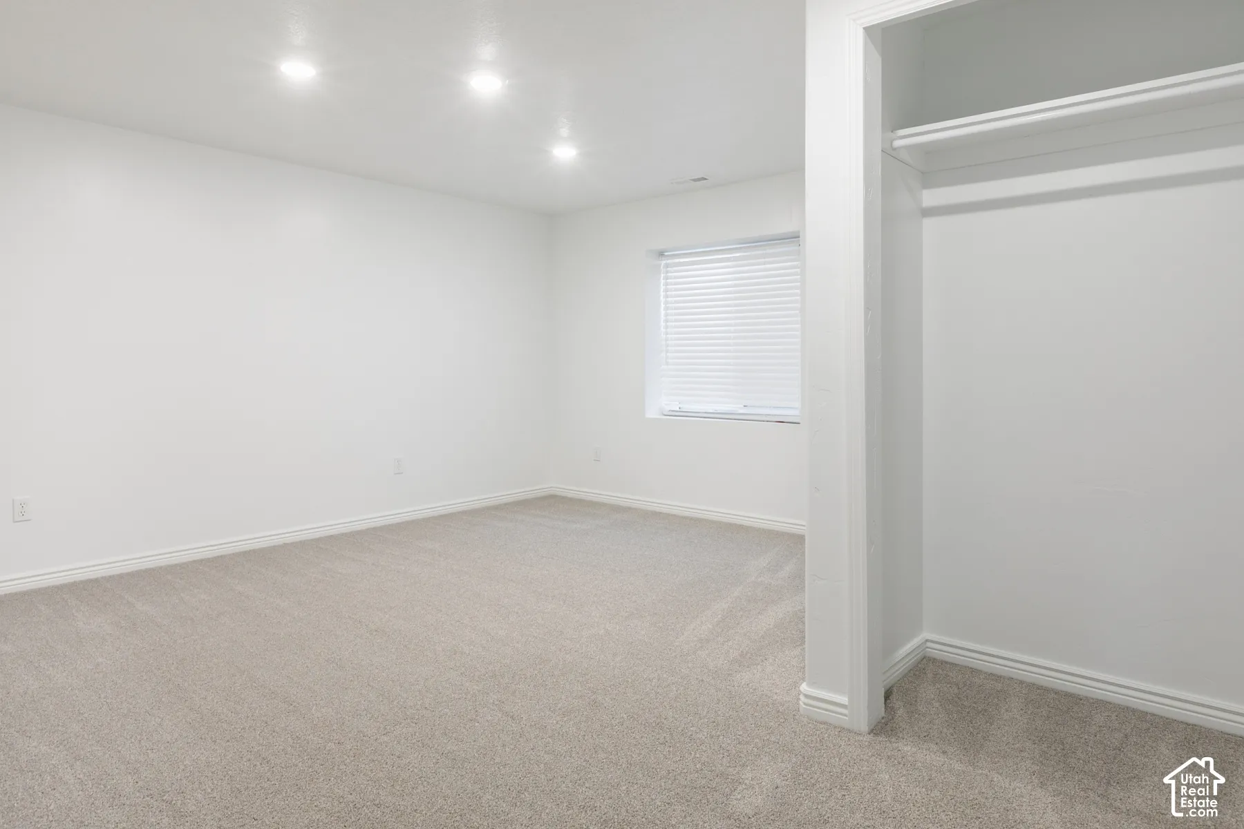 Recessed lighting illuminates a neutral carpeted room with a built-in closet and window with horizontal blinds