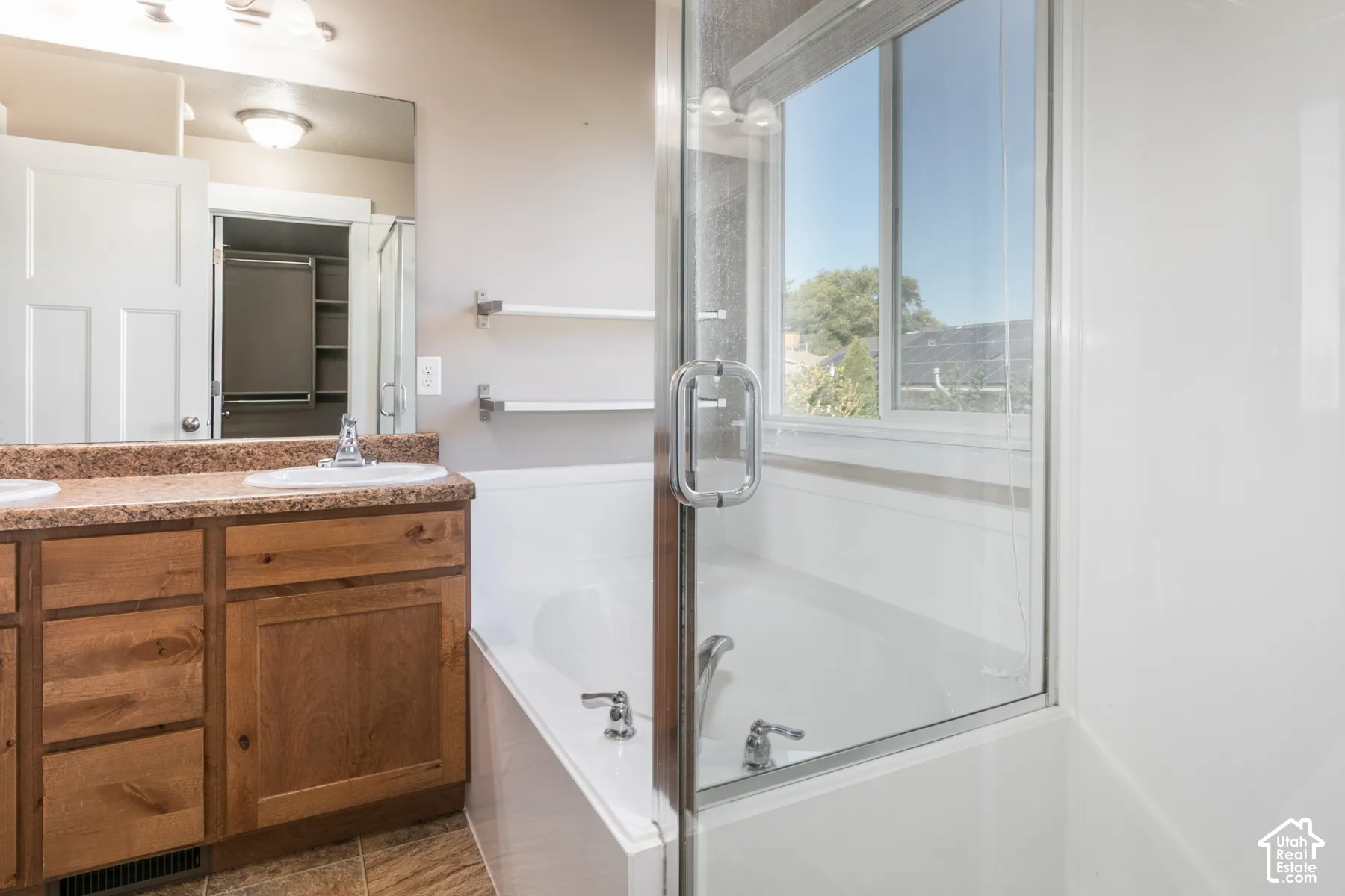 Dual sink vanity with wood-finish cabinetry, integrated bathtub with glass enclosure, and window