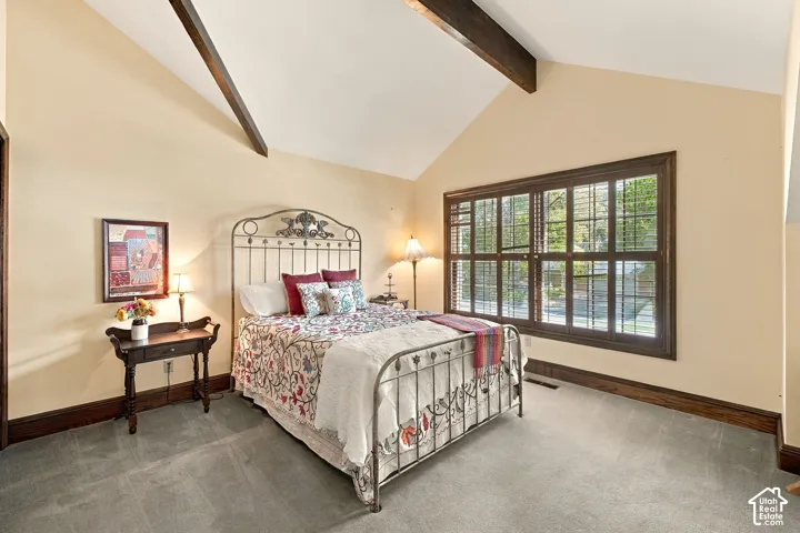Vaulted ceiling with exposed wood beams, expansive window with plantation shutters, dark wood trim, and wall-to-wall carpeting