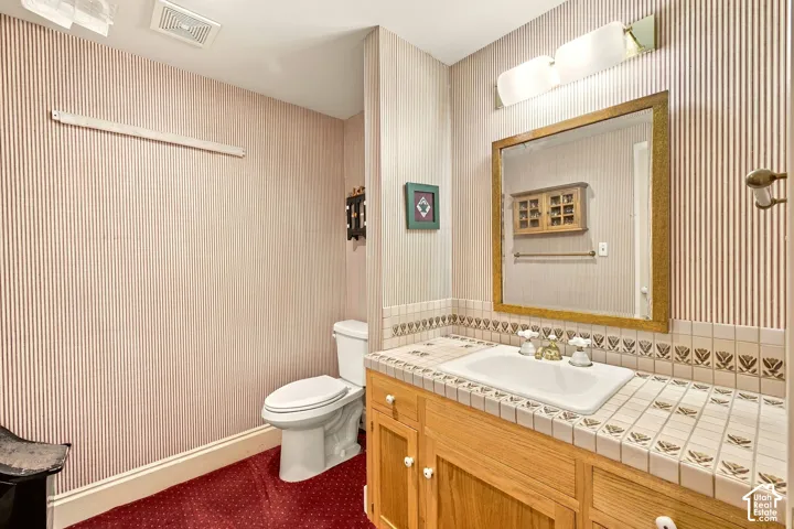 White ceramic toilet, integrated vanity sink, tiled countertop, wood-finish vanity cabinetry, and a framed wall mirror with overhead lighting