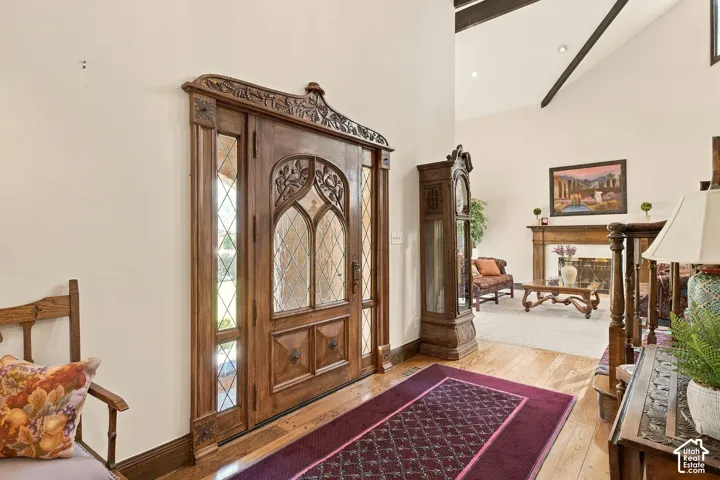 Ornate wooden entry door with leaded glass sidelights, wood flooring, vaulted ceiling with exposed beams, and a fireplace with a wood mantel