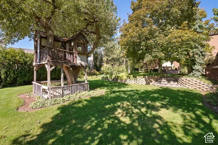 Expansive backyard featuring a manicured lawn, established shade trees, a multi-level treehouse with a wooden railing, and block retaining walls