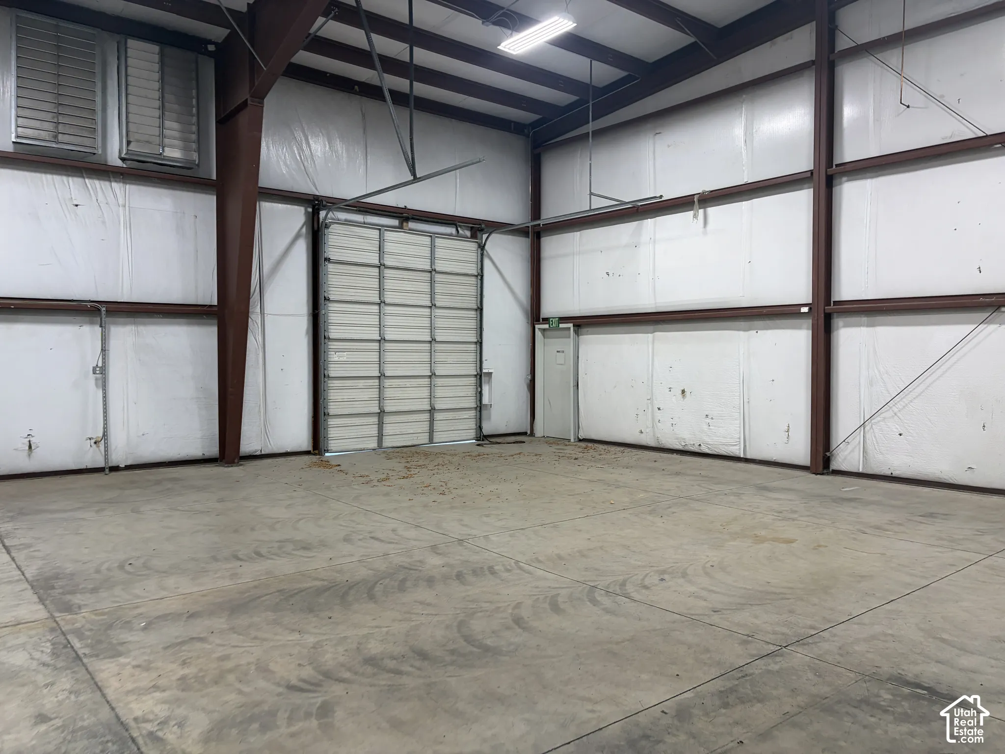 Concrete flooring, exposed structural beams, ceiling-mounted lighting, large roll-up door, and an additional entry door