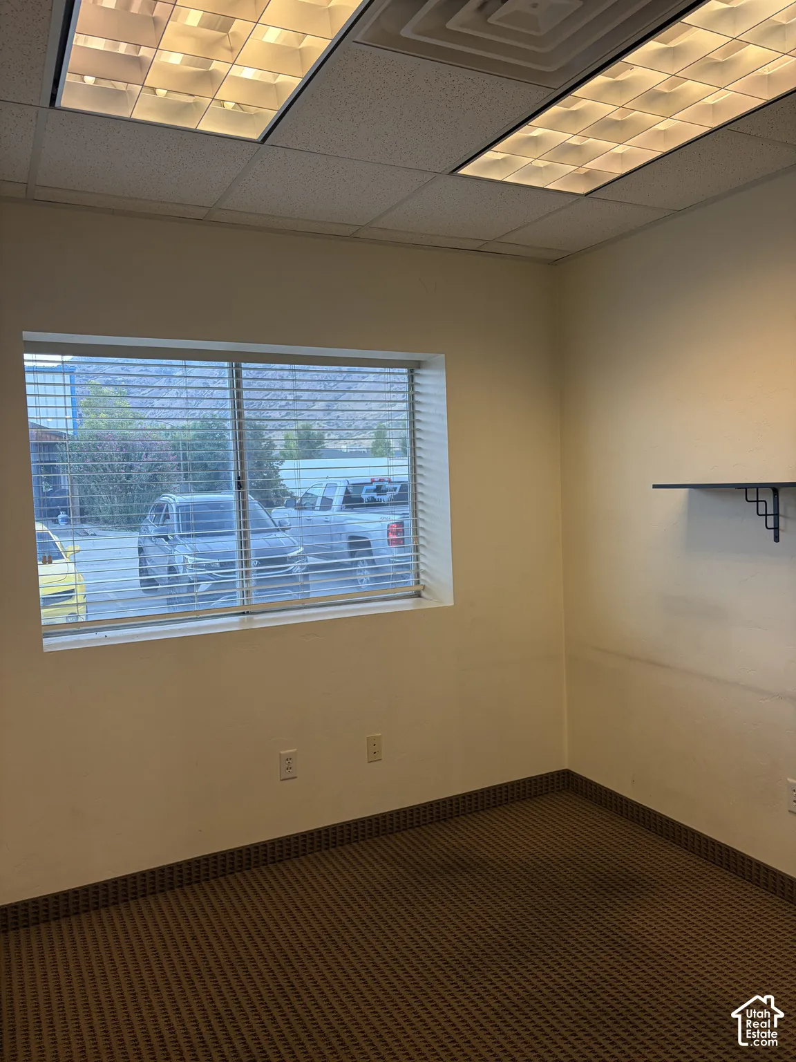 Window with horizontal blinds, drop ceiling with integrated lighting, wall-mounted shelf bracket, and patterned carpet flooring