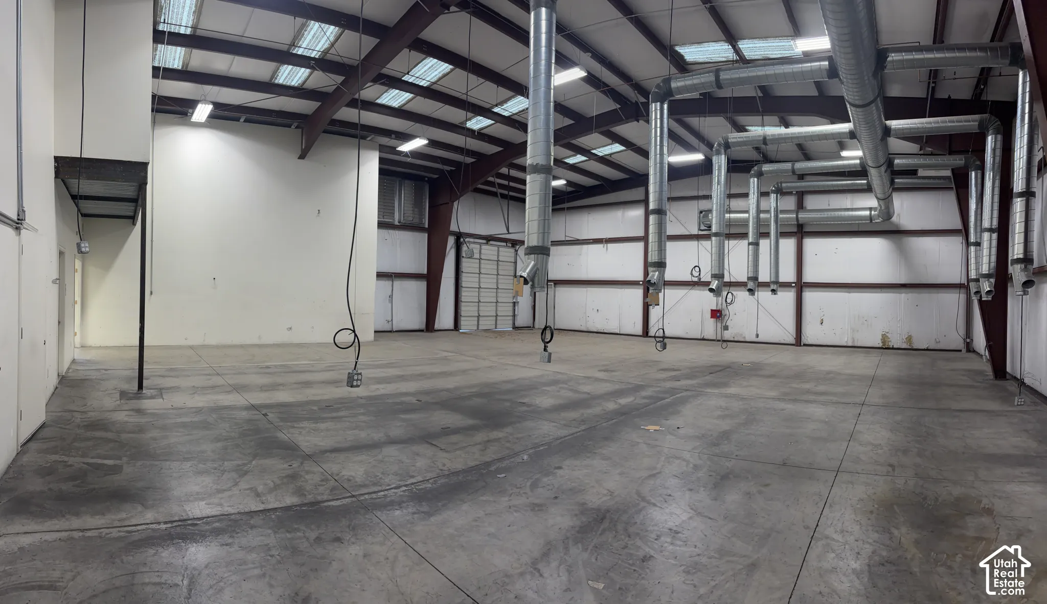 Expansive concrete flooring with high ceilings, featuring exposed metal trusses and extensive ductwork, complemented by multiple overhead skylights and bright overhead lighting