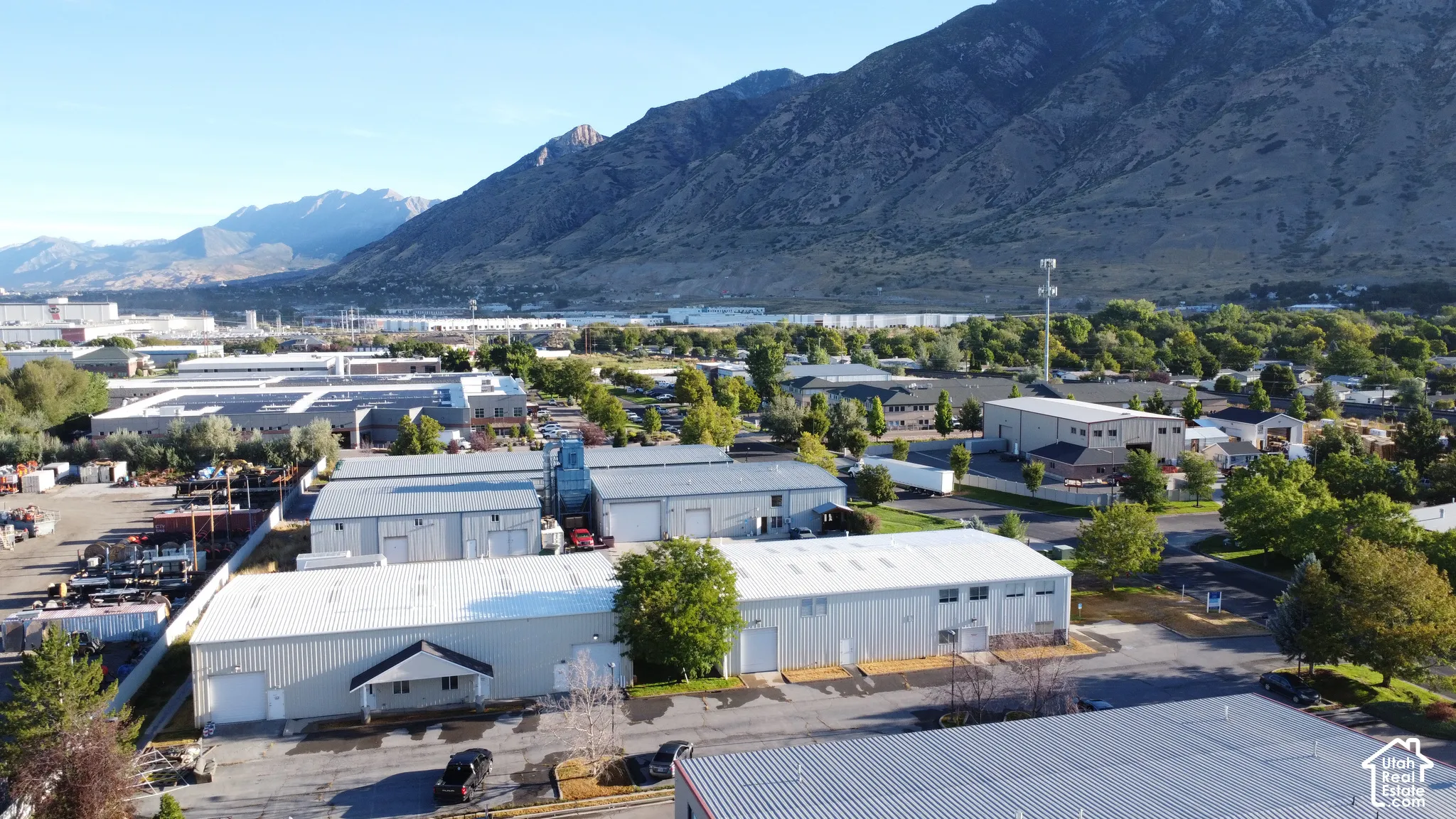 Industrial property featuring multiple buildings with metal siding and pitched roofs, surrounded by paved areas and mature trees, with a prominent mountain backdrop