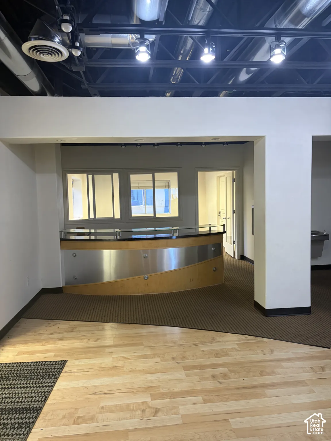 Hardwood flooring, exposed ceiling with ductwork and track lighting, curved reception desk with wood-finish and metal accents, interior windows, and a built-in water fountain