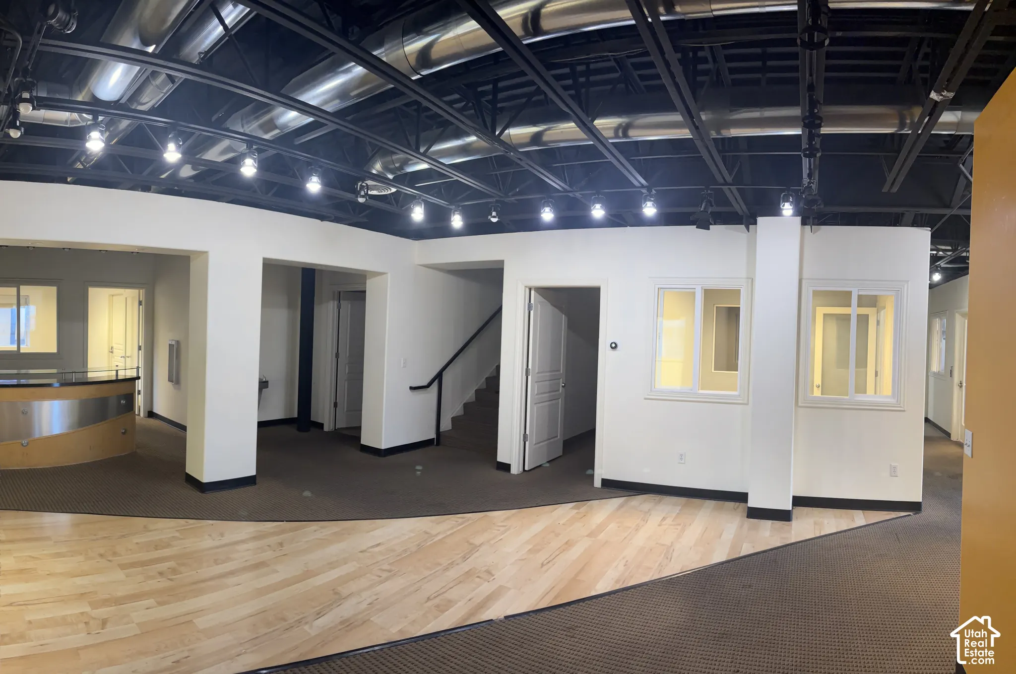 Open-concept commercial space featuring exposed ceiling ductwork, track lighting, a built-in reception desk with a wood-finish counter, and a staircase with a black handrail