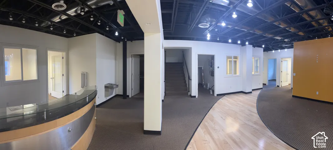 Open-concept space featuring a reception desk with a glass top and wood-finish base, exposed black ceiling with track lighting, mixed flooring with carpet and wood-finish sections, and a prominent staircase with a dark handrail