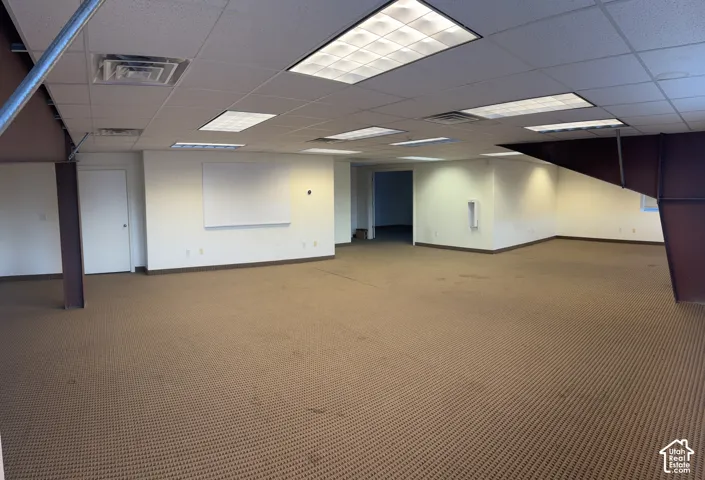 Expansive open area with patterned carpeting, drop ceiling with integrated lighting, and white wall finishes