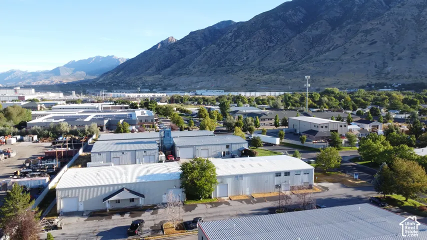 Industrial property featuring multiple buildings with metal siding and pitched roofs, surrounded by paved areas and mature trees, with a prominent mountain backdrop