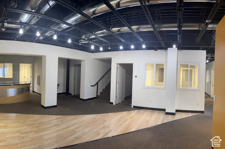 Open-concept commercial space featuring exposed ceiling ductwork, track lighting, a built-in reception desk with a wood-finish counter, and a staircase with a black handrail