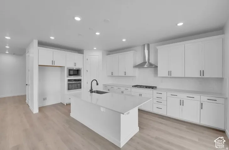 White shaker cabinetry with dark hardware, a center island with sink and faucet, integrated built-in appliances, a stainless steel range hood, and wood-finish flooring