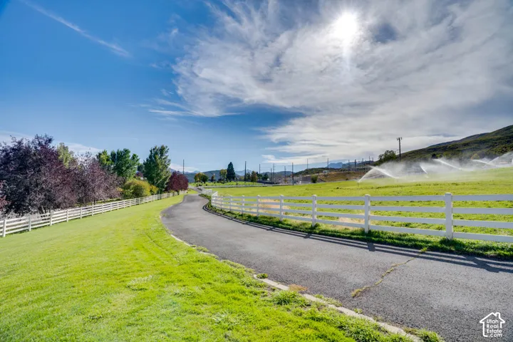 Paved driveway with white fencing, expansive green lawn, mature trees, and open fields with active irrigation