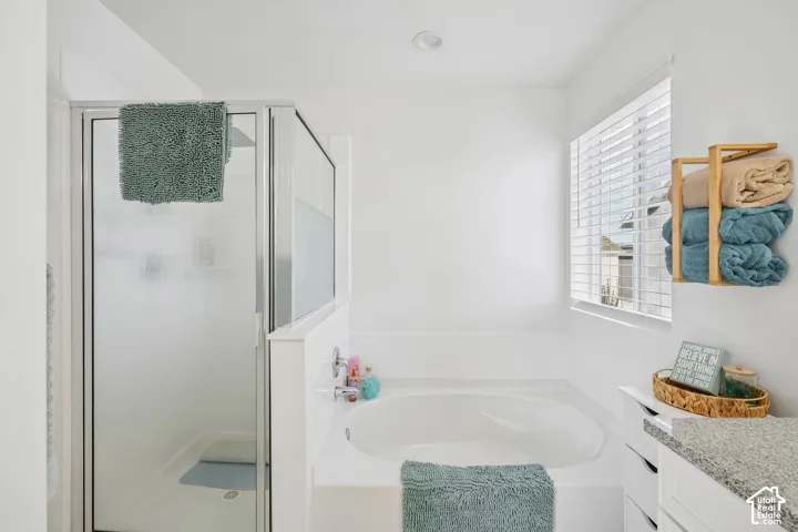 Walk-in shower with clear glass door, built-in soaking tub, window with blinds, and white cabinetry with a light-colored countertop