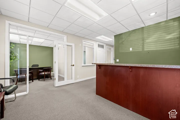 Wood-finish reception counter with a speckled countertop, neutral carpet flooring, drop ceiling, and French doors leading to an office space