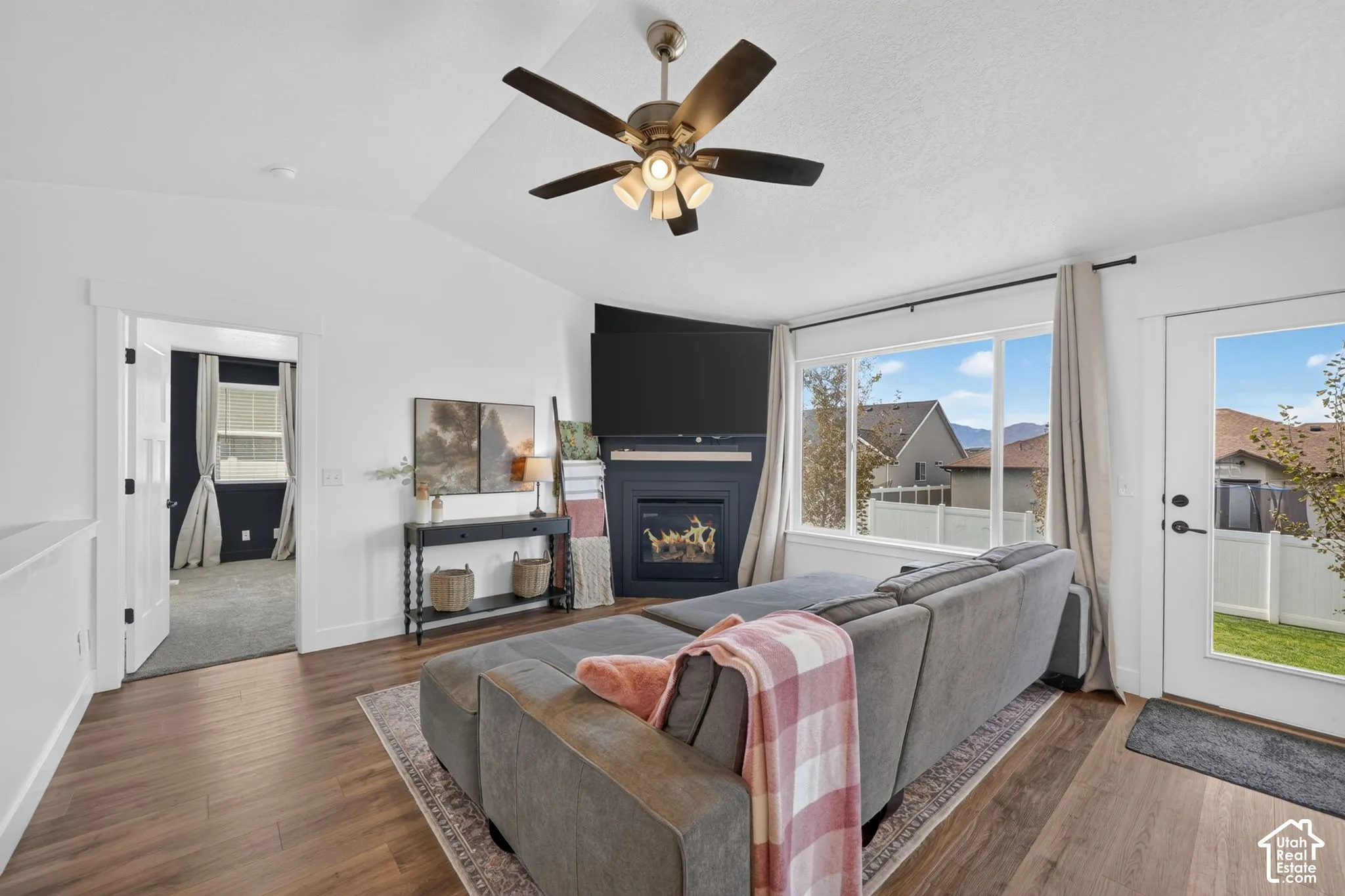 Living area featuring wood-finish flooring, an angled fireplace with built-in media space, large windows, a ceiling fan, and a glass-paneled exterior door
