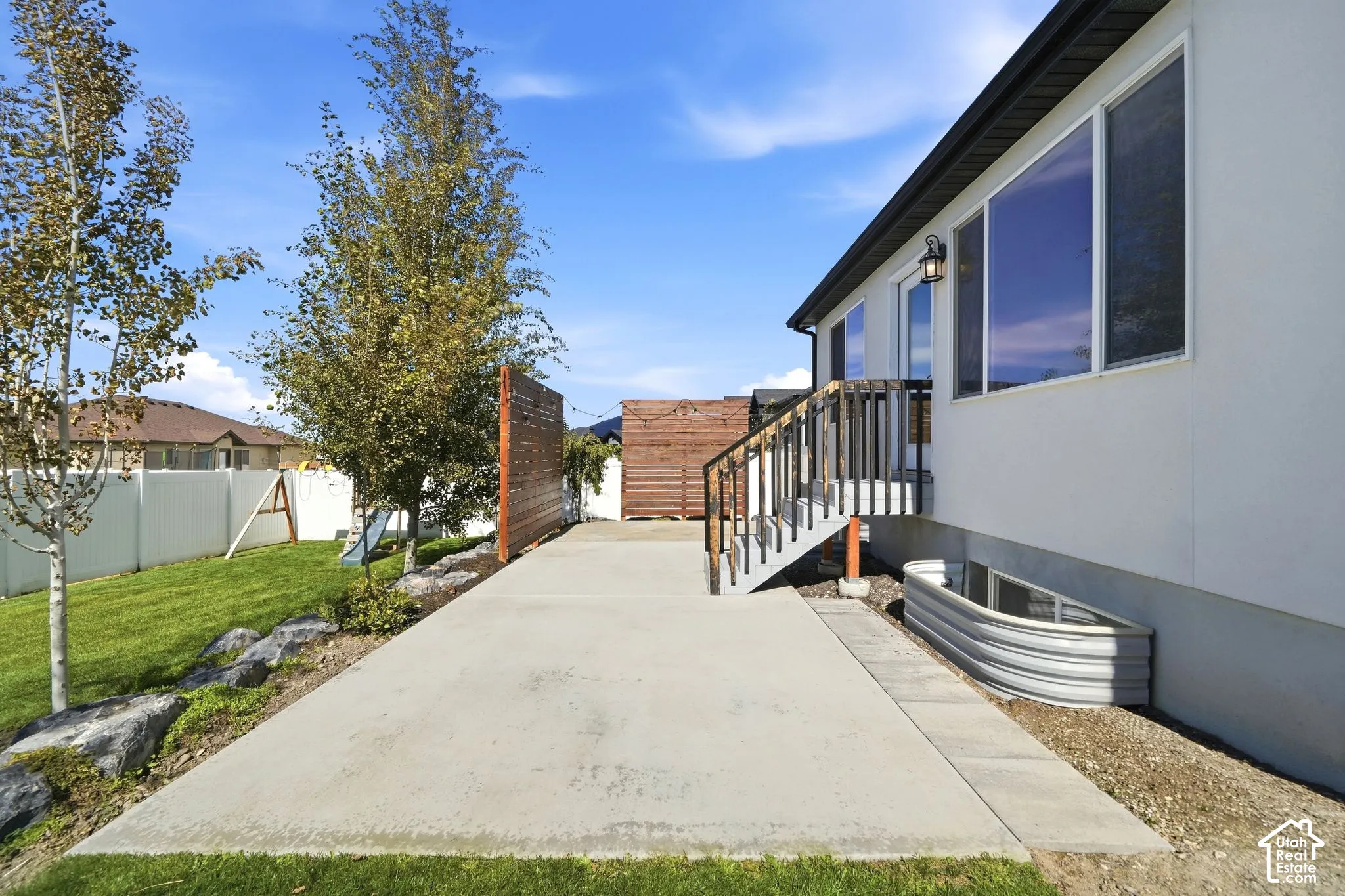 Concrete patio with adjacent grass area, exterior stairs with dark railing, a window well, and a privacy fence