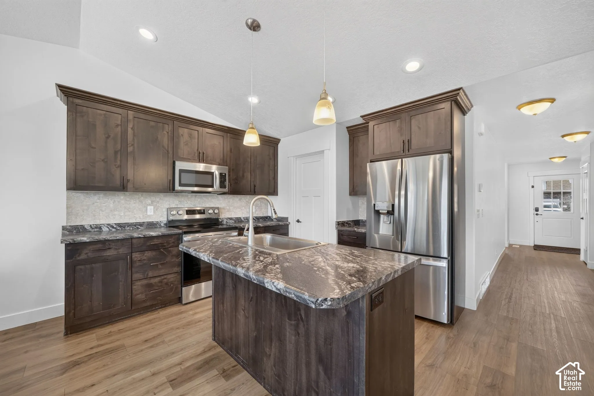 Kitchen featuring dark wood-finish cabinetry, stainless steel appliances, a center island with a sink, and pendant lighting