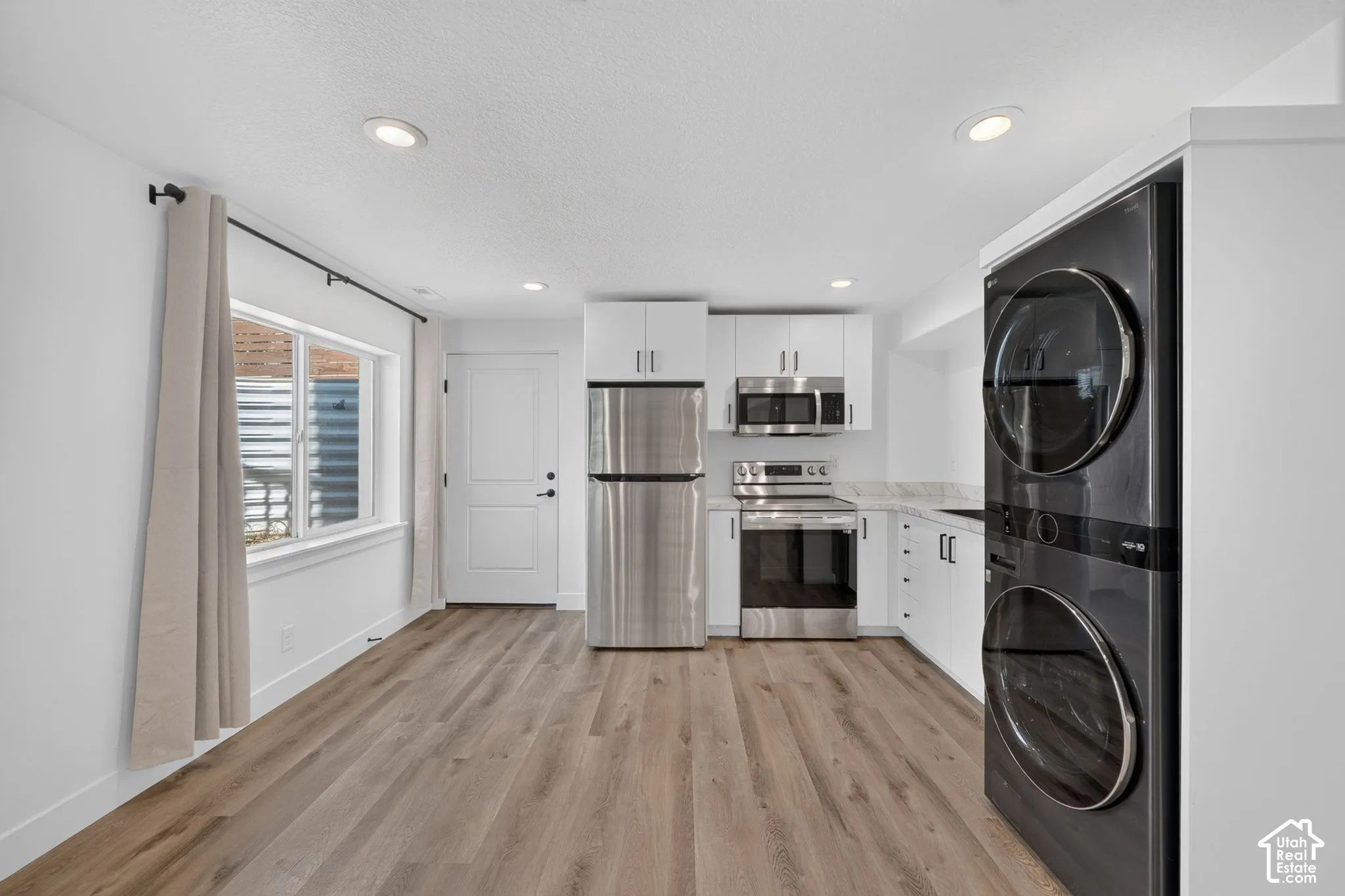 Wood-finish flooring, recessed lighting, stainless steel appliances, stacked laundry units, and white cabinetry