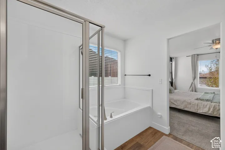 Walk-in shower with glass enclosure, white soaking tub with brushed nickel fixtures, wood-finish flooring, and window with blinds
