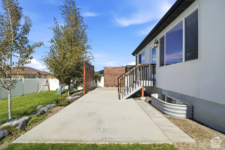 Concrete patio with adjacent grass area, exterior stairs with dark railing, a window well, and a privacy fence