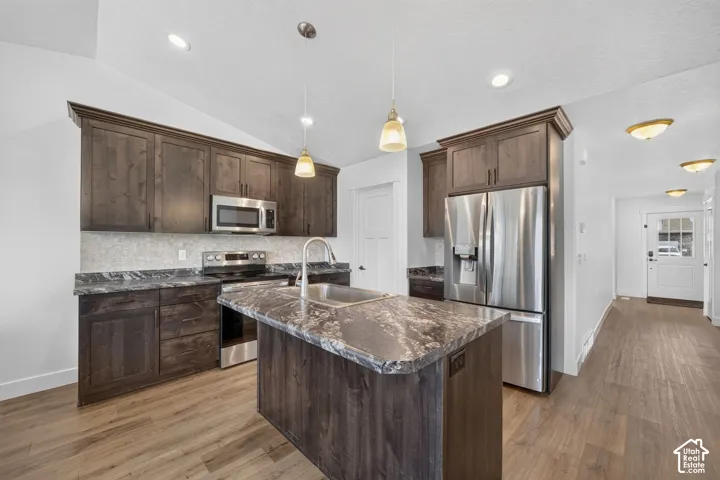 Kitchen featuring dark wood-finish cabinetry, stainless steel appliances, a center island with a sink, and pendant lighting