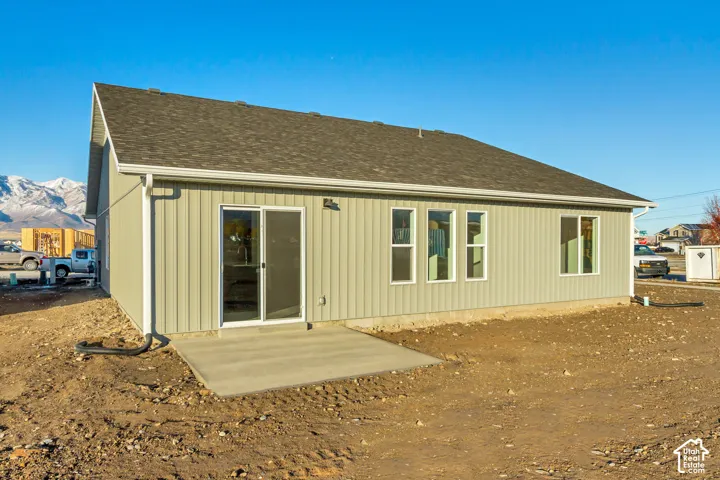 Newly constructed residence featuring vertical siding, a dark shingle roof, white gutters, multiple windows, and a concrete patio with a sliding glass door