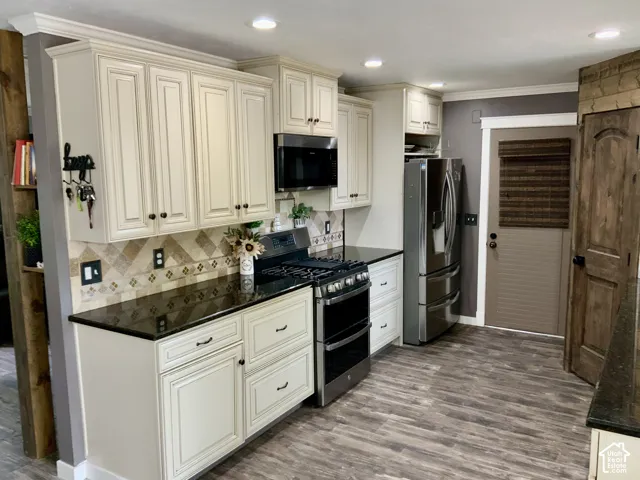 Cream-toned cabinetry, dark countertops, patterned tile backsplash, stainless steel appliances, and wood-finish flooring