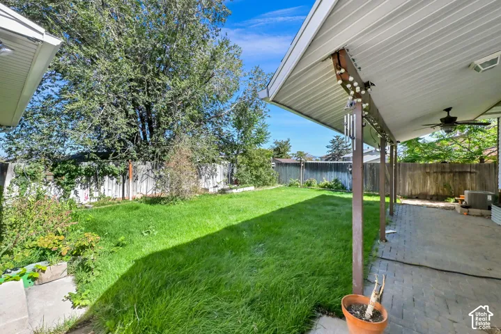 Expansive backyard featuring a green lawn, wood fencing, and a covered patio with a ceiling fan and paver flooring