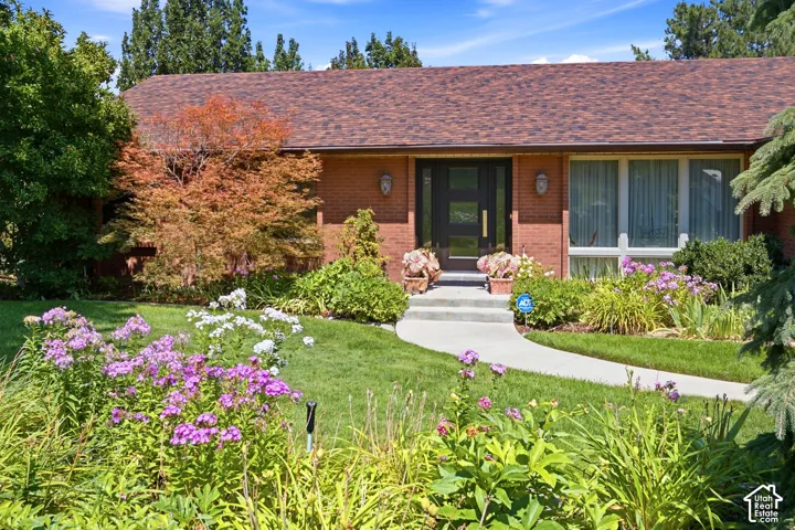 Brick exterior with a brown shingle roof, a front walkway, and a landscaped yard featuring mature trees and flowering plants