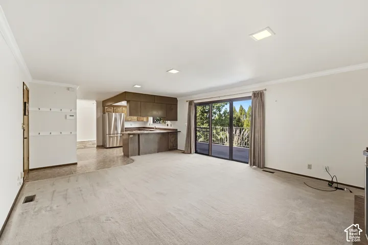 Open-concept living area with carpet flooring, crown molding, recessed lighting, and a kitchen featuring wood cabinetry, stainless steel refrigerator, and a breakfast bar