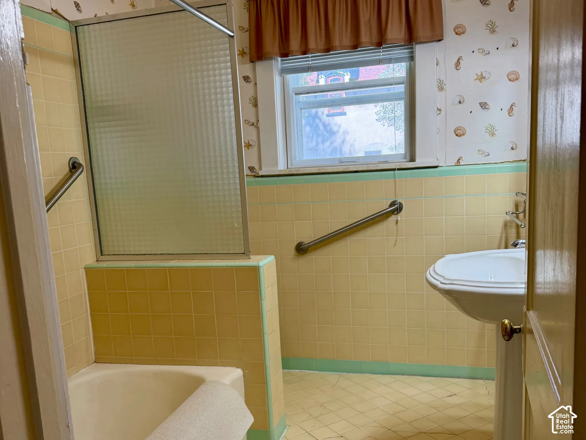 Pedestal sink, tiled walls, built-in bathtub, glass shower enclosure, and window with privacy blinds